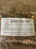 A clear plastic bag of The Herb Girls Orange Spice Tea filled with loose-leaf tea, resting on a rustic wooden surface. The label is white with black text and features the brand logo.